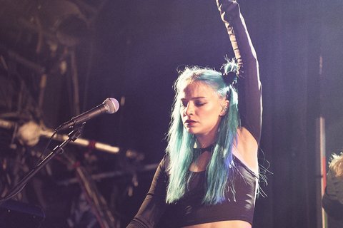 Eine Musikerin mit langen, türkisfarbenen Haaren steht an einem Mikrofon. Sie trägt ein schwarzes Oberteil und hebt einen Arm, während sie konzentriert auf die Musik blickt. Im Hintergrund ist eine dunkle Bühnenumgebung zu erkennen.