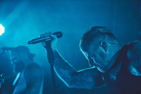 Zwei Musiker auf der Bühne in blauer Beleuchtung. Der vordere Sänger hält ein Mikrofon und hat eine konzentrierte Haltung. Der Hintergrund zeigt einen weiteren Musiker mit Gitarre. Die Atmosphäre ist energiegeladen und intensiv.