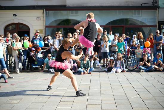 Quator Stomp Zwei Performer in pinken Tutus führen akrobatische Tänze auf einer Bürgersteigbühne auf, während eine große Menschenmenge zuschaut. Die Atmosphäre ist lebhaft und fröhlich, mit verschiedenen Zuschauern, die applaudieren und sich amüsieren.