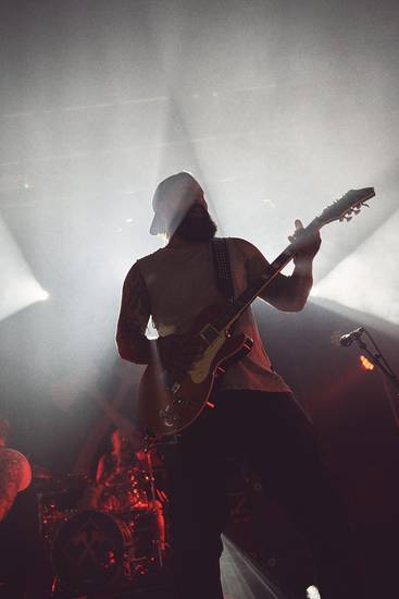 Ein Musiker spielt Gitarre auf der Bühne, umgeben von Lichtstrahlen. Die Silhouette des Spielers ist sichtbar, während im Hintergrund eine weitere Person und die Bühne angedeutet sind. Die Atmosphäre ist dynamisch und musikalisch.