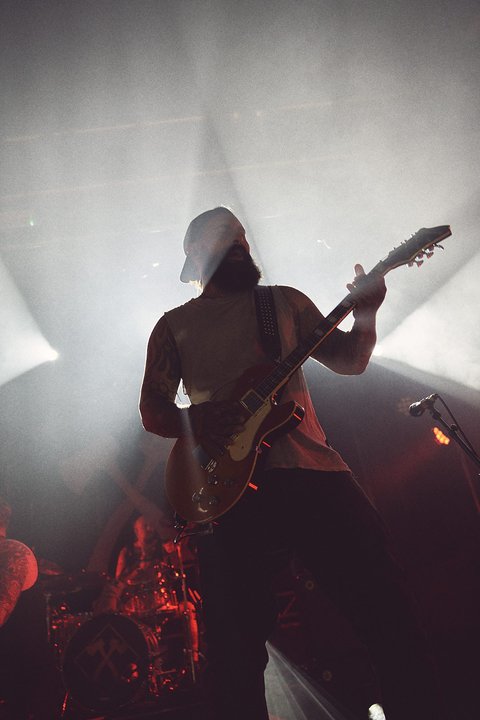 Ein Musiker spielt Gitarre auf der Bühne, umgeben von Lichtstrahlen. Die Silhouette des Spielers ist sichtbar, während im Hintergrund eine weitere Person und die Bühne angedeutet sind. Die Atmosphäre ist dynamisch und musikalisch.