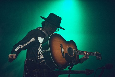 Ein Musiker spielt Gitarre auf der Bühne, gekleidet in ein schwarzes Oberteil mit Skelettmuster und einem breiten Hut. Der Hintergrund ist in grünliches Licht getaucht, was eine lebendige Konzertatmosphäre schafft.