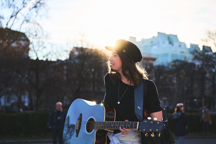Eine junge Frau mit Gitarre steht im Freien, umgeben von Bäumen und Menschen. Die Sonne scheint hinter ihr, wodurch sie in ein warmes Licht getaucht wird. Sie trägt ein schwarzes T-Shirt und einen Hut.