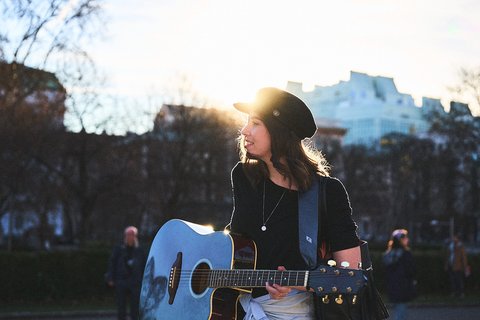 Eine junge Frau mit Gitarre steht im Freien, umgeben von Bäumen und Menschen. Die Sonne scheint hinter ihr, wodurch sie in ein warmes Licht getaucht wird. Sie trägt ein schwarzes T-Shirt und einen Hut.