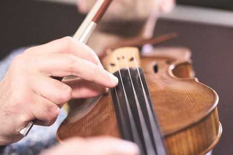 Ein Musiker spielt Violine, indem er mit einer Hand die Saiten drückt und mit dem Bogen in der anderen Hand über die Saiten streicht. Der Fokus liegt auf den Händen und der Violine, während der Musiker konzentriert aussieht.