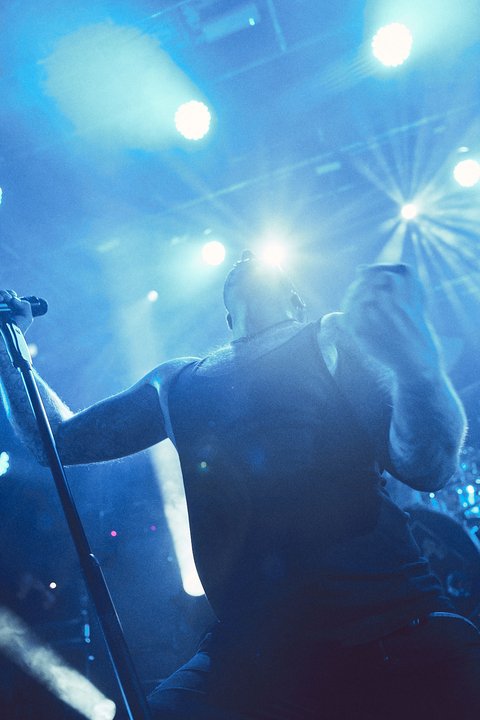 Ein Sänger steht auf der Bühne und hält ein Mikrofon in der Hand. Er ist stark beleuchtet von blauen und weißen Scheinwerfern, die eine lebendige Konzertatmosphäre erzeugen. Die Zuschauer sind im Hintergrund nicht sichtbar.