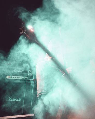Ein verschwommener Musiker steht auf einer Bühne, umgeben von grünlichem Nebel. Die Silhouette der Gitarre ist sichtbar, während im Hintergrund ein Verstärker zu erkennen ist. Die Atmosphäre vermittelt ein Gefühl von Live-Musik und Energie.