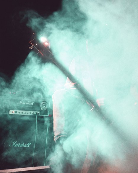 A guitarist is partially obscured by swirling green smoke on stage. The silhouette of the guitar and the speaker amplifier are visible, creating a mysterious and atmospheric musical setting. The image conveys a sense of energy and live performance.