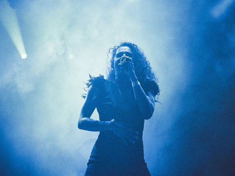 Eine Sängerin steht auf der Bühne und hält ein Mikrofon in der Hand. Sie trägt ein schwarzes Kleid und hat lockige Haare. Der Hintergrund ist in blauem Licht und Nebel gehüllt, was eine dramatische Atmosphäre schafft.