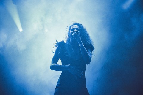 A singer with curly hair performs on stage, holding a microphone and expressing passion through her gestures. The atmosphere is dramatic, filled with blue smoke and soft lighting, creating an engaging concert ambiance.