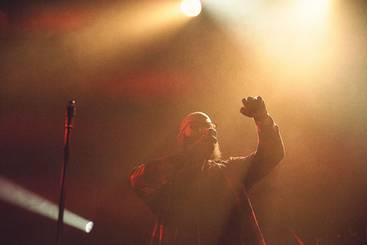 Faded Reality Ein Sänger steht auf der Bühne, umgeben von dramatischem Licht. Er hebt seine Hand, während er in das Mikrofon singt. Die Atmosphäre ist energiegeladen und erinnert an ein Live-Konzert.