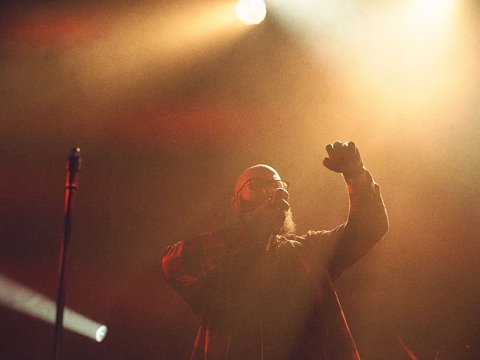 Ein Sänger steht auf der Bühne, umgeben von dramatischem Licht. Er hebt seine Hand, während er in das Mikrofon singt. Die Atmosphäre ist energiegeladen und erinnert an ein Live-Konzert.