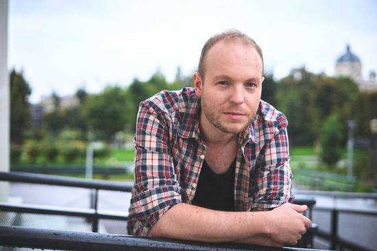 Ein junger Mann mit Kurzhaarschnitt und einem karierten Hemd lehnt entspannt auf einem Balkon. Im Hintergrund sind grüne Bäume und ein verschwommenes Stadtbild zu erkennen. Das Wetter scheint bewölkt zu sein.
