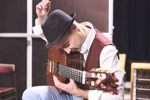 Ein Musiker sitzt auf einem Stuhl und spielt eine Gitarre. Er trägt einen schwarzen Hut und einen eleganten Anzug. Mit gebogenen Fingern zupft er die Saiten, während er sich konzentriert. Der Hintergrund ist unscharf, was den Fokus auf sein Spiel lenkt.