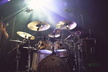 Ein Schlagzeug mit mehreren Toms, einem Snare und Becken, beleuchtet von Scheinwerfern. Die Umgebung deutet auf eine Live-Musik-Performance hin, mit Mikrofonen und dunklem Hintergrund.