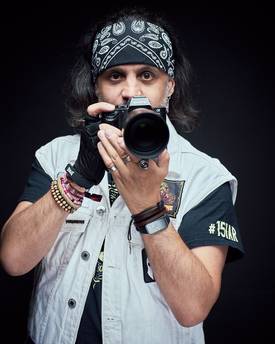 Ein Mann mit langen, dunklen Haaren trägt ein Bandana und ein ärmelloses Jackett. Er hält eine Kamera in der Hand und schaut direkt in die Linse. Auffällige Armbänder und ein T-Shirt mit einem grafischen Motiv ergänzen sein Outfit.