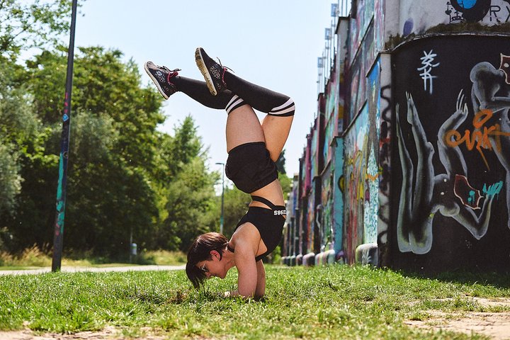 Eine Person steht kopfüber auf den Händen in einer sportlichen Pose auf einer Wiese. Im Hintergrund sind bunte Graffiti an einer Wand zu sehen. Der Himmel ist blau, und die Umgebung wirkt grün und lebendig.