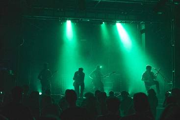 Eine Band spielt auf einer Bühne, umgeben von grünen Lichtern. Vier Musiker sind im Mittelpunkt, während ein Publikum im Vordergrund zu sehen ist. Die Atmosphäre ist lebhaft und energiegeladen.