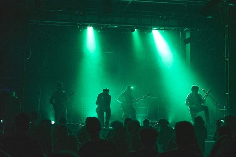 Eine Band spielt auf einer Bühne, umgeben von grünen Lichtern. Vier Musiker sind im Mittelpunkt, während ein Publikum im Vordergrund zu sehen ist. Die Atmosphäre ist lebhaft und energiegeladen.