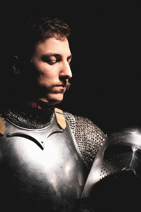 Ein Mann in einer Rüstung aus Kettenhemd und Plattenpanzer steht im Halbdunkel. Er schaut nachdenklich auf einen Helm, den er in der Hand hält. Sein Gesicht ist ernst und er hat einen kleinen Schnurrbart. Die Beleuchtung betont die Details seiner Rüstung.