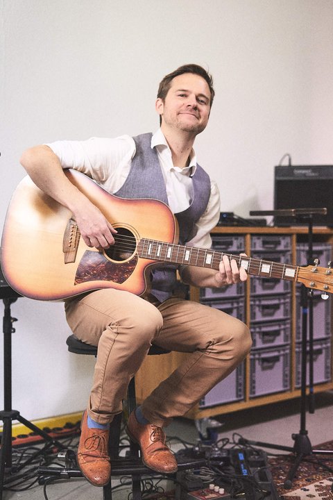 Ein Mann sitzt auf einem Stuhl und hält eine Akustikgitarre. Er trägt eine helle Weste und lächelt. Die Umgebung wirkt kreativ, mit Musikgeräten im Hintergrund. Der Fokus liegt auf seiner Freude am Musizieren.