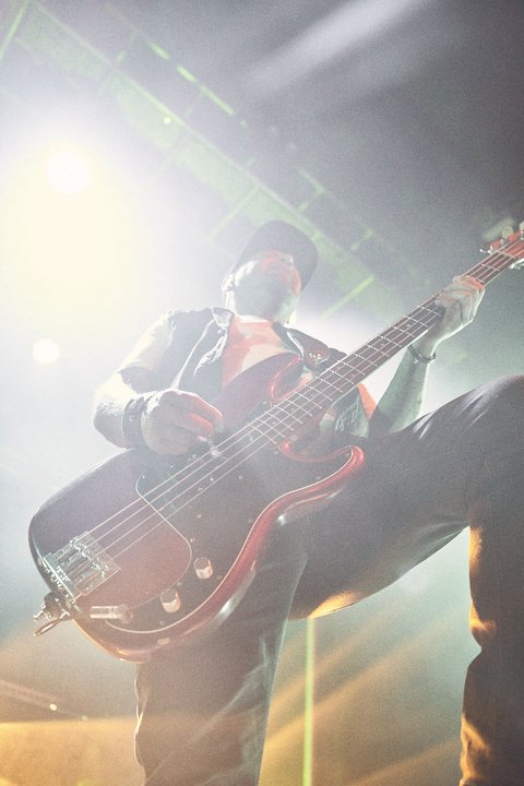 Ein Musiker spielt Bass auf einer Bühne. Er steht in einem Lichtstrahl und ist energiegeladen, während er sein Instrument präsentiert. Die Atmosphäre scheint dynamisch und lebhaft zu sein, was auf ein live Konzert hindeutet.