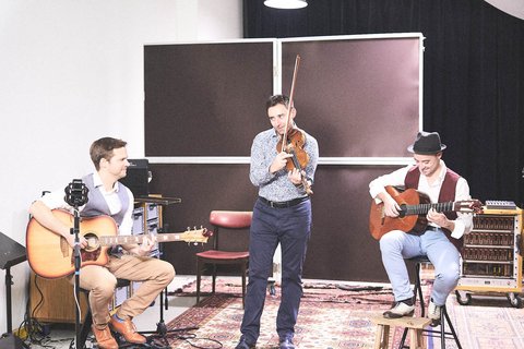 Drei Musiker stehen in einem Raum: ein Mann mit einer akustischen Gitarre, ein anderer mit einer Violine, und ein dritter sitzt mit einer klassischen Gitarre. Sie spielen gemeinsam Musik und wirken konzentriert. Im Hintergrund sind einige Instrumente und eine gemütliche Teppichfläche zu sehen.
