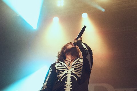 Ein Sänger steht auf der Bühne, trägt ein Outfit mit Skelett-Design und hält ein Mikrofon. Er ist in einer dynamischen Pose abgebildet, während Lichter in verschiedenen Farben hinter ihm leuchten. Die Atmosphäre ist energiegeladen, passend zu einem Live-Auftritt.