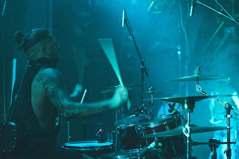 Ein männlicher Schlagzeuger mit Tattoos spielt leidenschaftlich an einem Schlagzeug. Er trägt ein ärmelloses Oberteil und ist von blauen Lichtern umgeben, die eine dynamische Atmosphäre erzeugen. Rauch ist im Hintergrund sichtbar, während er sich intensiv auf seine Musik konzentriert.