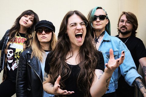 A group of five musicians poses together, exuding confidence and energy. The front woman, with long hair, gestures passionately while singing. The others stand behind her, showcasing diverse styles including leather jackets and sunglasses, creating a vibrant rock band vibe.