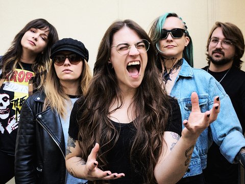 Fünf Musikerinnen und Musiker stehen zusammen vor einer neutralen Wand. Eine Sängerin in der Mitte schreit energetisch, während die anderen sie unterstützen und pose. Alle tragen lässige Kleidung, darunter Lederjacken und T-Shirts, was die rockige Atmosphäre unterstreicht.