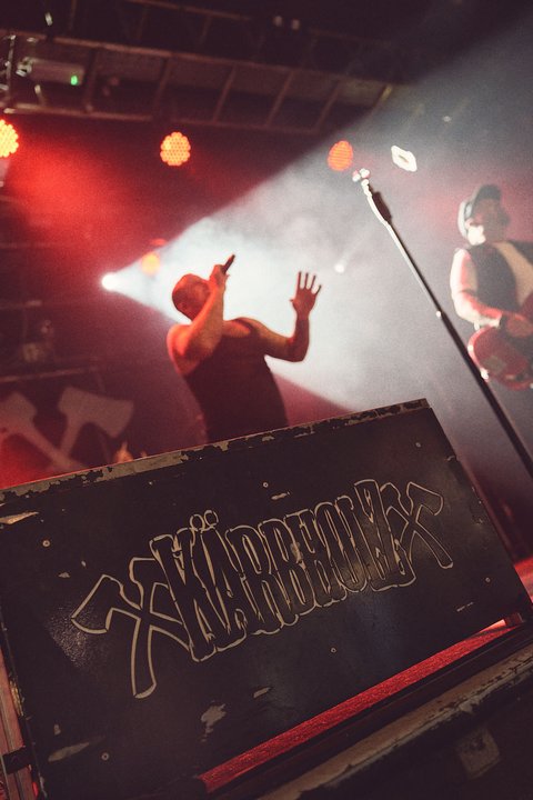 Ein Sänger steht auf einer Bühne und singt mit Ausdruck. Im Hintergrund sind unscharfe Silhouetten von weiteren Musikern und Lichtern zu sehen. Die Band hat ein auffälliges Logo auf dem Bühnenaufbau. Die Atmosphäre ist energetisch und lebhaft.