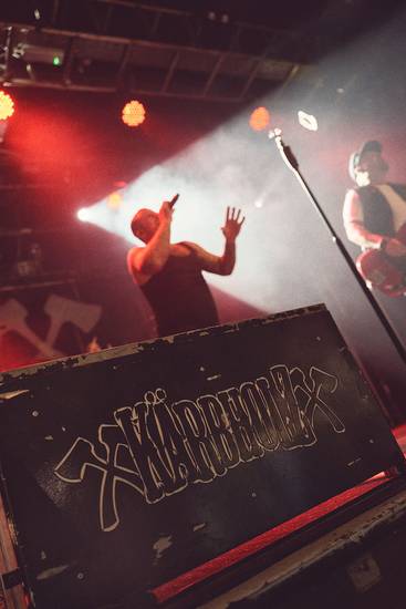 Ein Sänger steht auf einer Bühne und singt mit Ausdruck. Im Hintergrund sind unscharfe Silhouetten von weiteren Musikern und Lichtern zu sehen. Die Band hat ein auffälliges Logo auf dem Bühnenaufbau. Die Atmosphäre ist energetisch und lebhaft.