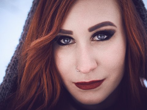 Porträt einer Frau mit langen, roten Haaren und auffälligem Make-up. Sie hat leuchtende Augen und einen direkten, ansprechenden Blick. Ihr Lippenstift ist in einem dunklen Ton gehalten. Die Umgebung hat einen frostigen, winterlichen Charakter.