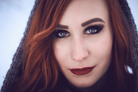 A close-up portrait of a woman with long, flowing red hair. She has striking blue eyes and bold makeup, including dark eyeliner and deep red lipstick. The background features a soft, blurred winter landscape, emphasizing her features.