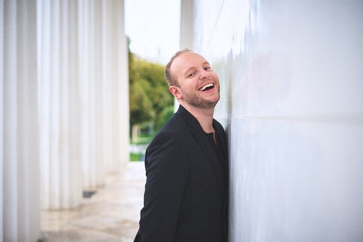Ein Mann in einem schwarzen Anzug lächelt fröhlich und lehnt sich an eine helle Wand. Im Hintergrund sind Säulen und eine grüne Landschaft sichtbar. Die Szene vermittelt eine entspannte und positive Atmosphäre.