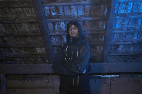 Ein junger Mann steht in einem dunklen, schattigen Raum unter einem Holzdach. Er trägt einen Kapuzenpullover und hat eine nachdenkliche Ausdrucksweise. Das Licht hat einen bläulichen Farbton, der die Atmosphäre geheimnisvoll wirken lässt.