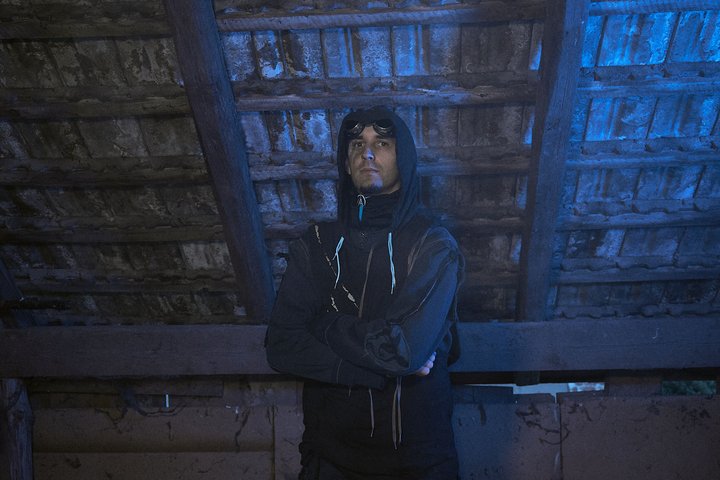 d0ck3r Ein junger Mann steht in einem dunklen, schattigen Raum unter einem Holzdach. Er trägt einen Kapuzenpullover und hat eine nachdenkliche Ausdrucksweise. Das Licht hat einen bläulichen Farbton, der die Atmosphäre geheimnisvoll wirken lässt.