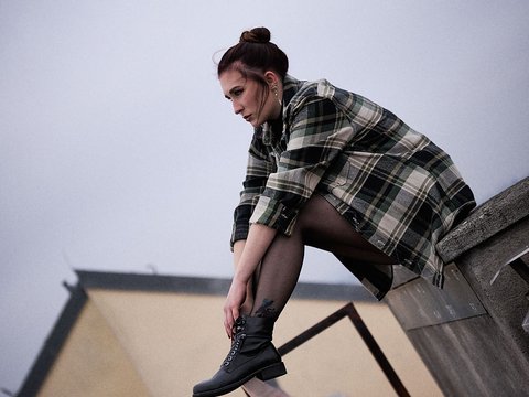 Eine junge Frau sitzt auf einer Mauer und blickt nach vorne. Sie trägt ein kariertes Hemd und schwarze Stiefel. Die Wolken am Himmel sind grau, was eine nachdenkliche Stimmung erzeugt. Die Umgebung wirkt urban und modern.