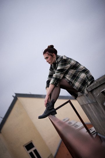 Eine junge Frau sitzt auf einer Mauer und blickt nach vorne. Sie trägt ein kariertes Hemd und schwarze Stiefel. Die Wolken am Himmel sind grau, was eine nachdenkliche Stimmung erzeugt. Die Umgebung wirkt urban und modern.
