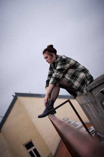 Eine junge Frau sitzt auf einer Mauer und blickt nach vorne. Sie trägt ein kariertes Hemd und schwarze Stiefel. Die Wolken am Himmel sind grau, was eine nachdenkliche Stimmung erzeugt. Die Umgebung wirkt urban und modern.
