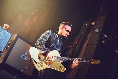 Ein Musiker spielt Gitarrenmusik auf der Bühne. Er trägt eine schwarze Lederjacke mit einem auffälligen Muster und Sonnenbrille. Im Hintergrund sind Lautsprecher und das Bühnenlicht sichtbar.