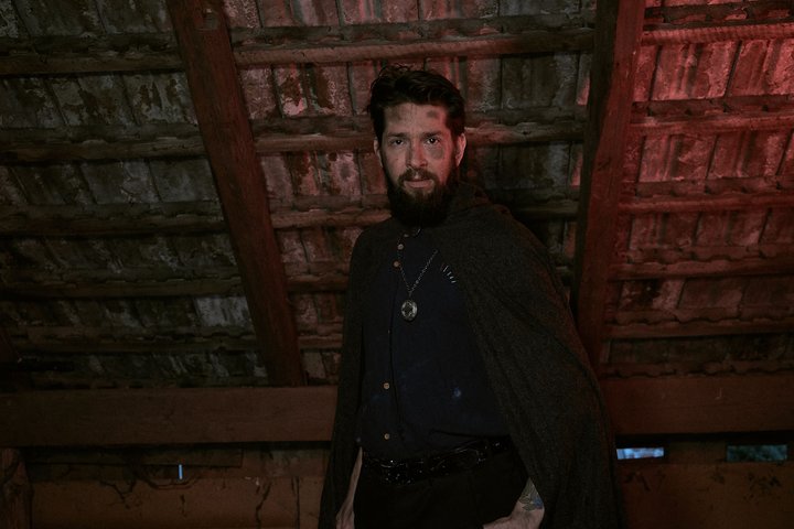 The GrandMaester Ein Mann mit Bart und einem Umhang steht in einem düsteren Raum mit einem gewölbten Dach. Er trägt eine Kette um den Hals und schaut direkt in die Kamera. Die Beleuchtung ist schwach und erzeugt eine geheimnisvolle Atmosphäre.