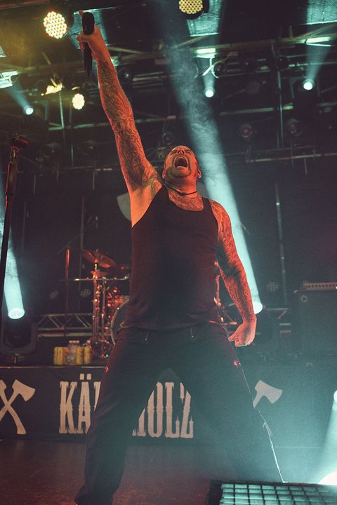 Ein Sänger hebt die Hand in die Luft und zeigt Begeisterung während eines Live-Auftritts. Er trägt ein schwarzes Tanktop und hat sichtbare Tattoos. Die Bühne ist von bunten Lichtern beleuchtet, die eine energetische Atmosphäre schaffen.