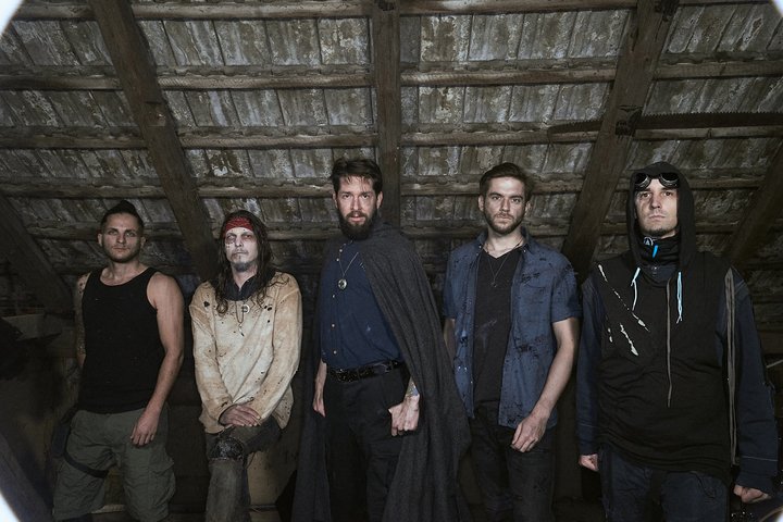 Aeons of Ashes Fünf Männer stehen in einem Raum mit Holzdecke. Sie tragen verschiedene Outfits, darunter eine schwarze Umhang und lässige Kleidung. Ihre Gesichtsausdrücke sind ernst, und sie scheinen gemeinsam in die Kamera zu blicken. Der Raum wirkt düster und erinnert an eine verlassene Umgebung.