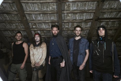 Fünf Männer stehen in einem Raum mit Holzdecke. Sie tragen verschiedene Outfits, darunter eine schwarze Umhang und lässige Kleidung. Ihre Gesichtsausdrücke sind ernst, und sie scheinen gemeinsam in die Kamera zu blicken. Der Raum wirkt düster und erinnert an eine verlassene Umgebung.