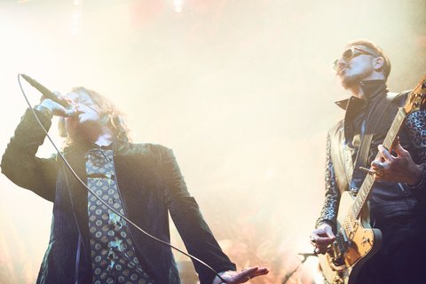 Zwei Musiker stehen auf einer Bühne in einem Konzert, einer singt ins Mikrofon, während der andere Gitarre spielt. Die Szene ist von buntem Licht und einem kreativen Nebel umgeben, was eine lebendige Atmosphäre erzeugt.