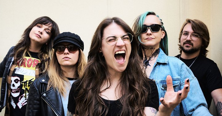 Eine Rockband mit fünf Mitgliedern posiert vor einer neutralen Wand. Die Sängerin in der Mitte zeigt eine kraftvolle Geste und wirkt energisch. Die anderen Bandmitglieder um sie herum tragen lässige Kleidung und lächeln oder schauen ernst. Die Szene strahlt Selbstbewusstsein und Gemeinschaft aus.