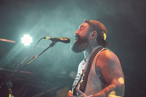 Ein Musiker mit Bart und Cap steht auf der Bühne und singt in ein Mikrofon. Er greift eine E-Gitarre und ist umgeben von stimmungsvollem Licht und Nebel. Die Darstellung vermittelt eine energiegeladene Live-Performance.