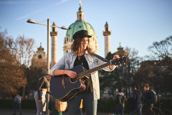 Eine junge Frau mit Gitarre steht vor einer historischen Architektur. Sie trägt eine Jacke und eine Mütze, während sie spielt und sich auf ihr Instrument konzentriert. Im Hintergrund sind Bäume und weitere Personen, die sich im Freien aufhalten, sichtbar.